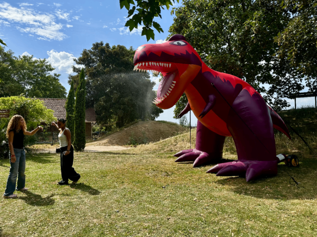 Dinosaure brumisateur gonflable de dos avec sa queue pointue qui arrose deux personnes
