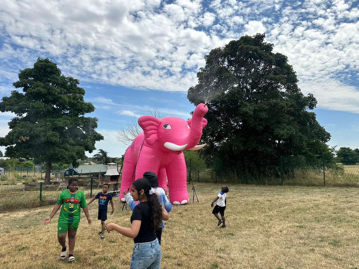 un éléphant Rose Gonflable brumisateur qui envoie de l'eau par la trompe avec un ciel bleu et des enfants qui se rafraichissent en dessous