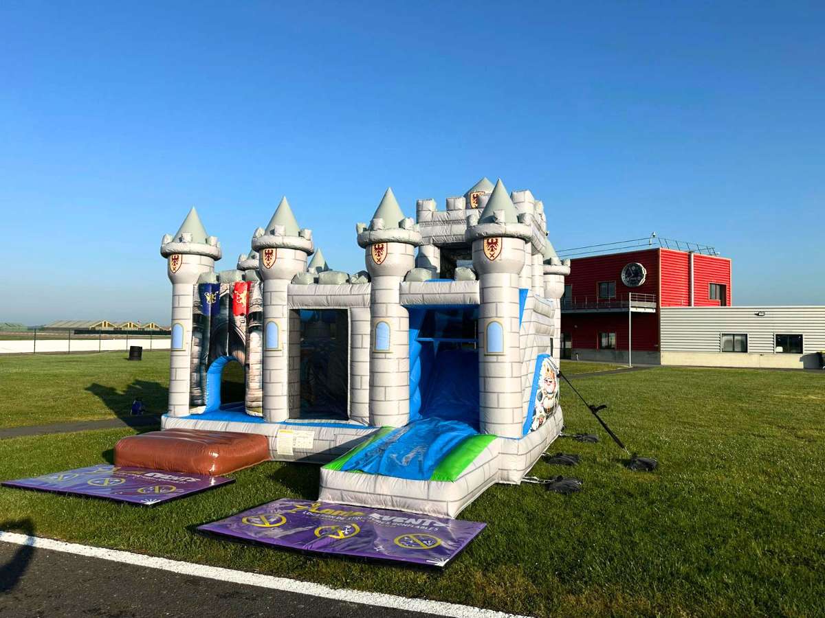 Château médiéval  gris gonflable avec plusieurs tours sur un terrain avec des enfants qui joue autour surveillé par un adulte