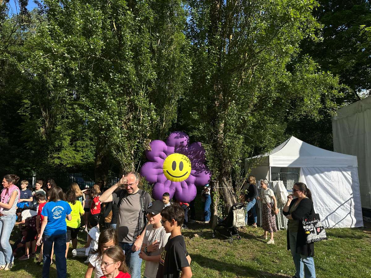 fleur gonflable violette avec smiley jaune suspendu entre deux arbres à un mètre du sol avec du monde autour