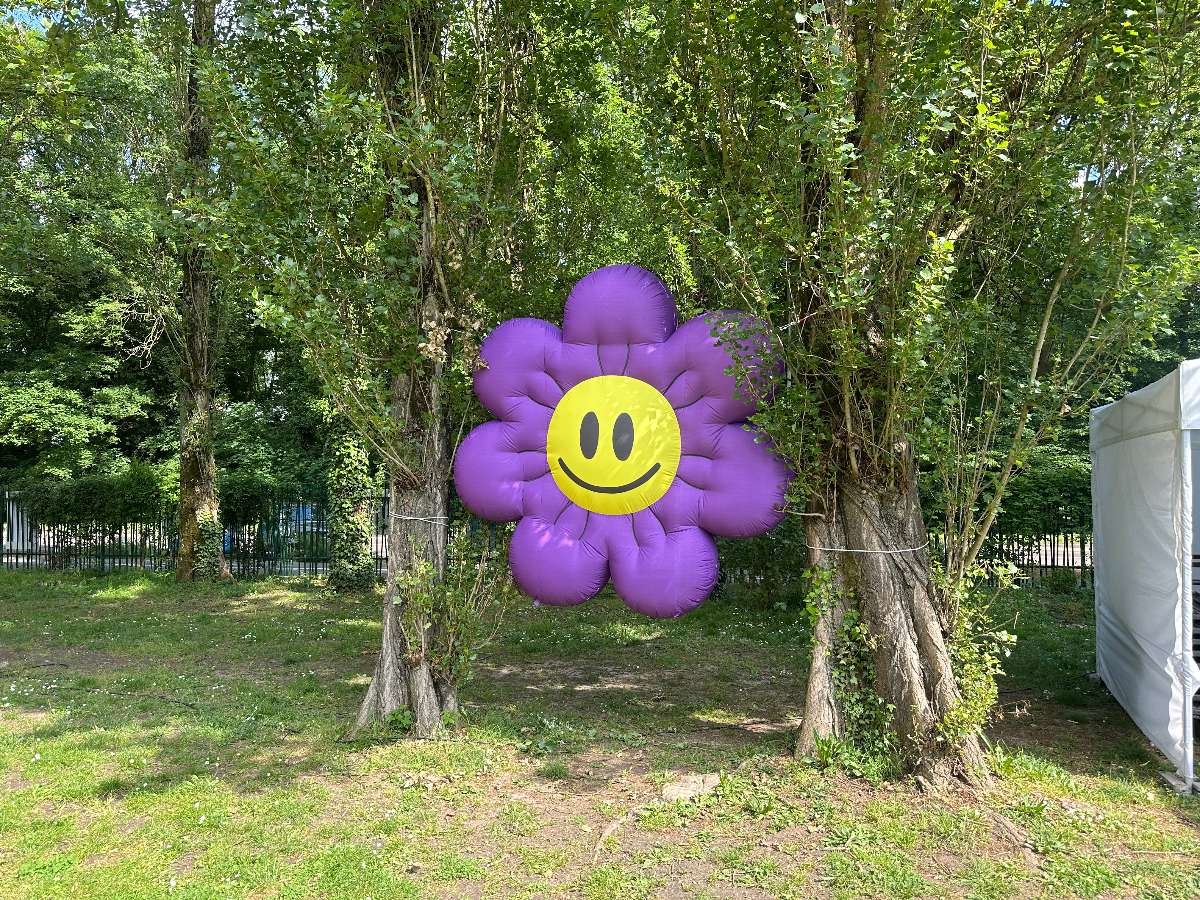 fleur gonflable violette avec smiley jaune suspendu entre deux arbres à un mètre du sol vue de face