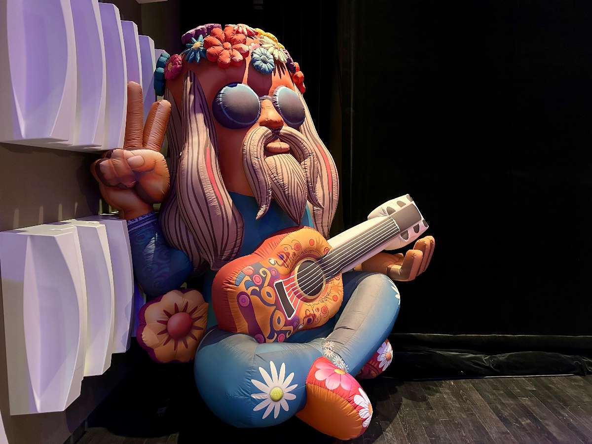 Guitariste hippie gonflable avec une guitare orange  et les doigts en peace vue de biais dans une pièce sombre 