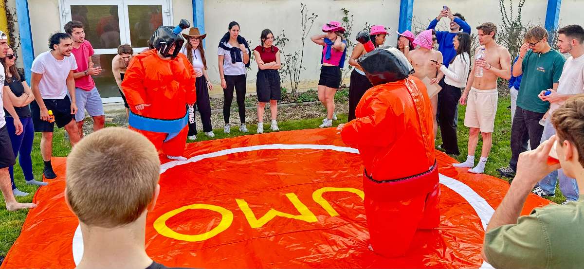 un combat de sumo adulte  en costume orange avec du monde autour qui observe  