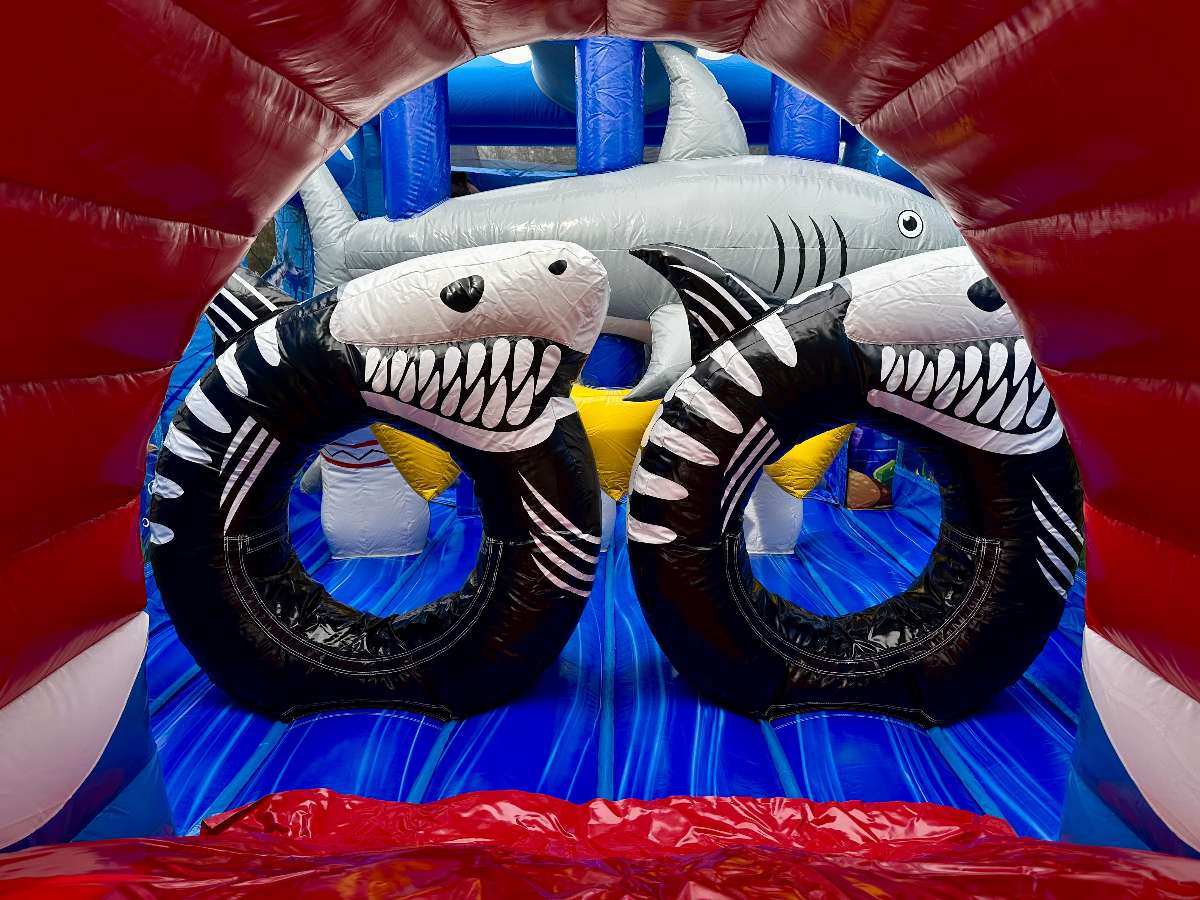 Vue intérieure d’un parcours d’obstacles gonflable thème océan avec structure bleue, obstacles souples en forme de petits requins gris, zone d’escalade et filets de sécurité. Décor marin imprimé avec poissons, espace de jeu ludique et sécurisé pour enfants, parfait pour animations et événements.