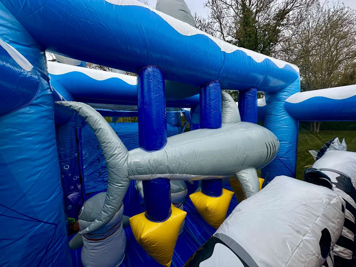 Vue intérieure d’un parcours d’obstacles gonflable thème océan avec structure bleue, obstacles souples en forme de petits requins gris, zone d’escalade et filets de sécurité. Décor marin imprimé avec poissons, espace de jeu ludique et sécurisé pour enfants, parfait pour animations et événements.